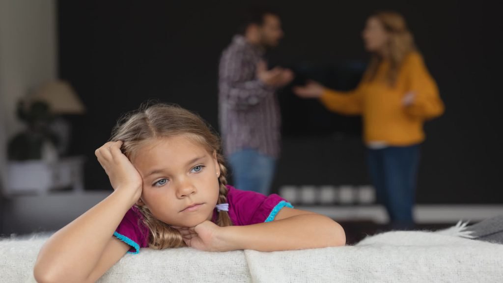 A young girl sitting sadly while her parents argue in the background